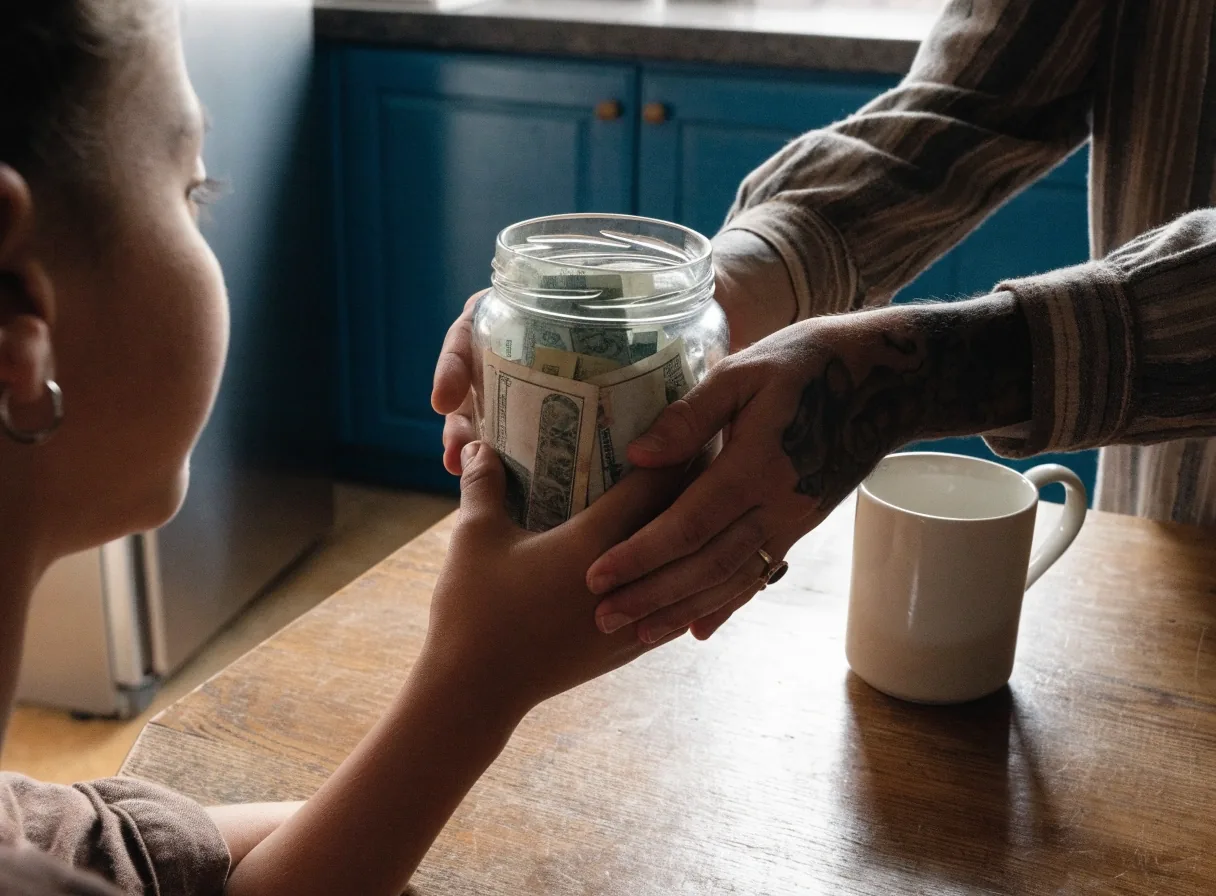 Mesa de cocina a la tarde con un frasco de ahorro, un cuaderno abierto y manos adultas ordenando dinero